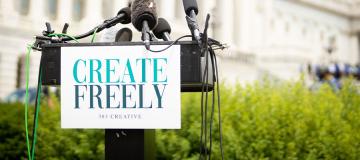 Microphones sit on a podium with a sign that reads 'Create Freely' in front of the U.S. Capitol